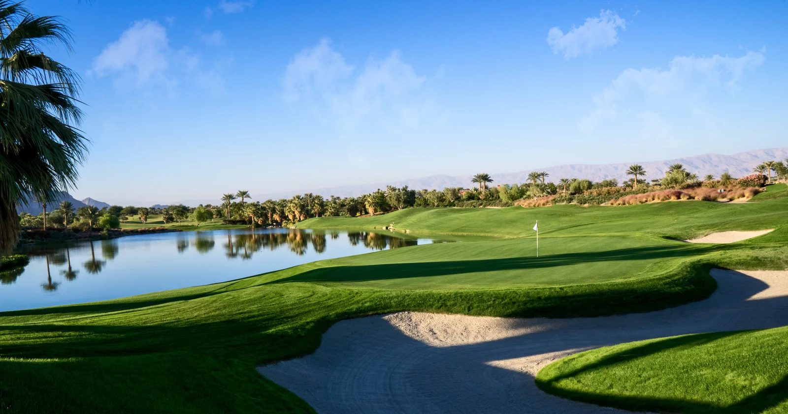 Ein Golfplatz bei untergehender Sonne gesäumt von Palmen. In der Mitte ist ein klarer großer Teich zu sehen. Im Hintergrund lassen sich Hügel erahnen.